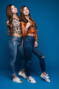 Two pretty slim sisters wearing checkered shirts, jeans and sneakers posing together on blue isolated background. Female sporty teens looking at camera and smiling. Concept of trends and style.