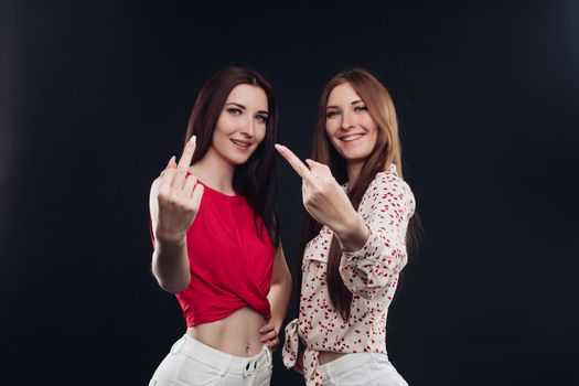 Two young girls in casual clothes gesturing by their fingers. Beautiful sisters standing close to each other and showing signs by hands. Brunette twins with long hair posing together at camera.