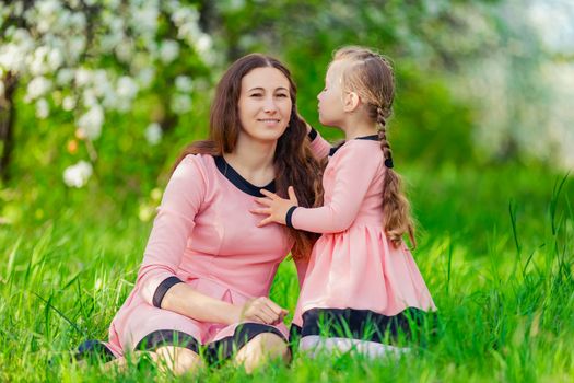 mother and daughter sit in the green grass against the backdrop of blooming apple trees