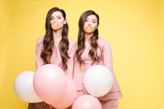 Young twins in elegant dresses holding pink and white balloons in their hands. Brunette sisters with long hair blowing big bubbles from chewing gum. Beautiful girls posing at camera with decorations.