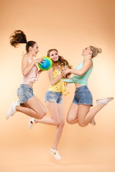 Group of happy young girls in casual clothes jumping high and getting fun. Beautiful women in colorful t-shirts smiling and playing with ball. Pretty models in jeans shorts spending holidays together.
