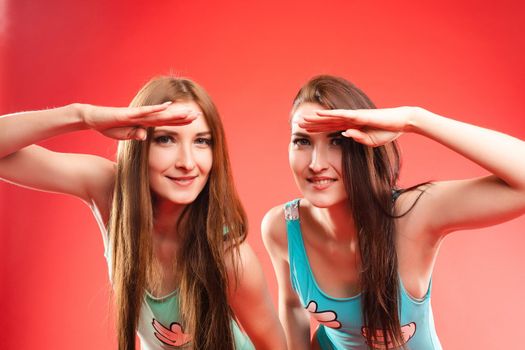 Front view of two athletic girl in swimming wear looking at camera and smiling on red isolated camera. Sporty women keeping hands near heads and looking for someone. Concept of energy and fitness.