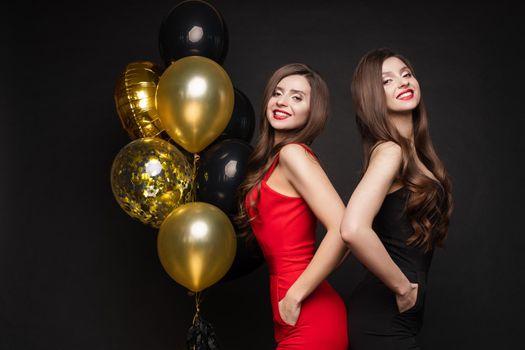 Cropped view of two beautiful sisters wearing red and black dresses and standing back to back in studio. Young long haired model posing near golden balloons, looking at camera and smiling on isolated.
