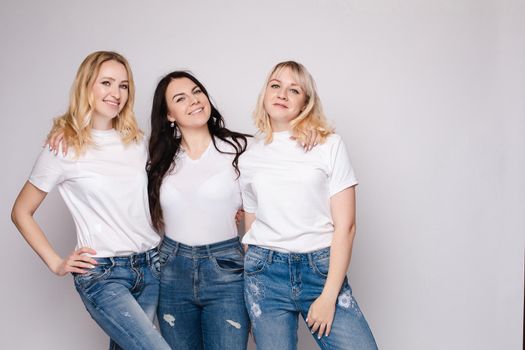 Front view of three beautiful women wearing white shirts and jeans and posing on isolated background. Stylish models standing in studio, looking at camera and smiling. Concept of casual street outfit.