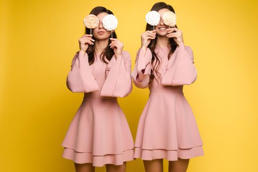 Front view of charming twins in pink dresses closing eyes with lollipops and posing on isolated background. Two female friends laughing and having fun in studio. Concept of childhood and beauty.