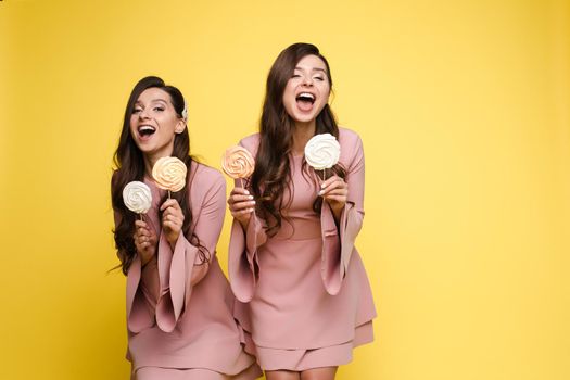 Front view of charming twins in pink dresses closing eyes with lollipops and posing on isolated background. Two female friends laughing and having fun in studio. Concept of childhood and beauty.