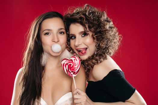 Front view of two different girls looking at camera and posing on black isolated background. Sexy brunette chewing gum while female friend laughing and keeping lollipop in hand. Concept of party.