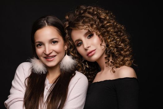 Portrait of two beautiful glamour girls in black and white standing together. Young model with curly hair leaning on her friend's shoulder. Happy pretty lady with pink feather earrings smiling shyly.