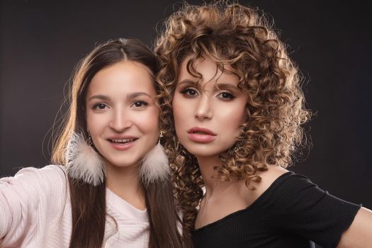 Portrait of two beautiful glamour girls in black and white standing together. Young model with curly hair leaning on her friend's shoulder. Happy pretty lady with pink feather earrings smiling shyly.