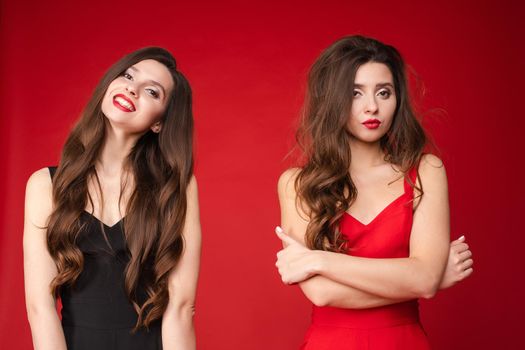 Front view of two different twins in black and red outfits looking at camera and posing on isolated background. Happy beautiful woman standing near sad shaggy sister with folded arms in studio.