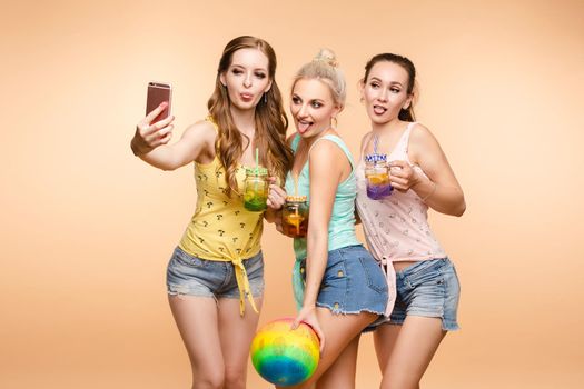 Studio portrait of stunning young girls in bright tops and denim shorts taking self-portrait with smartphone holding fresh homemade non-alcoholic drinks in special colored jars with straws. Isolate on yellow.