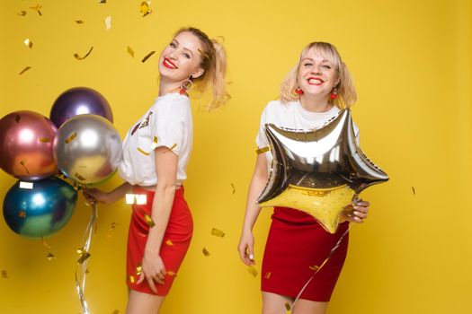 Two happy beautiful woman friend celebrating posing surrounded by colorful air balloon confetti. Attractive smiling female wearing bright clothes enjoying party isolated at yellow studio background