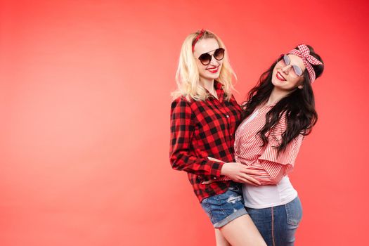 Studio shot of pretty smiling blonde and brunette girlfriends in sunglasses and trendy outlooks hugging against bright red background. Isolated.