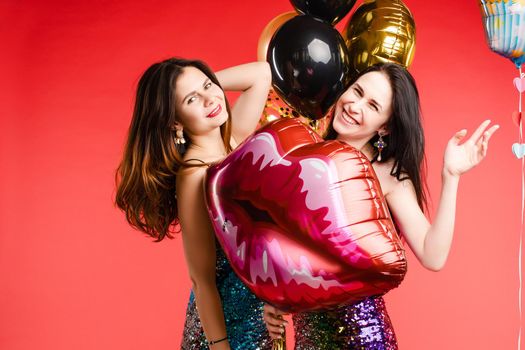 Close up of brunette twins with long hair in black and red costumes posing at camera. Young sisters standing together and holding red lips balloon. Gorgeous models with red lips sending air kisses.