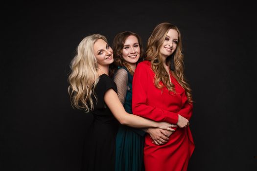 Studio full length portrait of three stunning young ladies in evening gowns posing for camera. Black background. New Year evening celebration birthday concept.