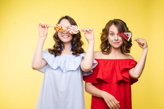 Studio portrait of gorgeous brunette sisters posing in trendy elegant dresses. Stylish outlooks. Smiling at camera. Yellow background with candy.