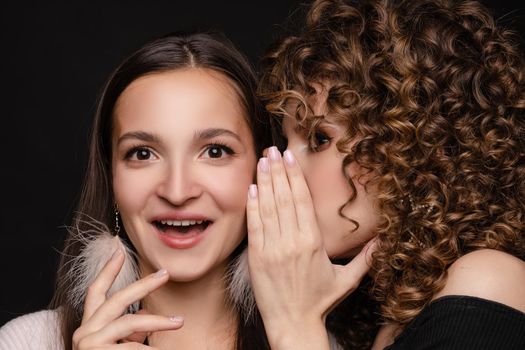 Front view of shocked brunette with big eyes and open mouth in white dress looking at camera while female friend whispering secret on black isolated background. Young women gossiping in studio.