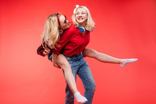 Studio shot of pretty smiling blonde and brunette girlfriends in sunglasses and trendy outlooks hugging against bright red background. Isolated.
