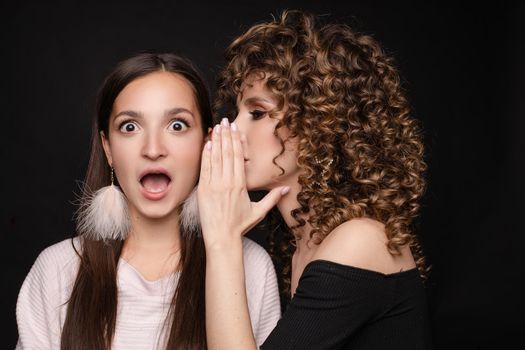 Front view of shocked brunette with big eyes and open mouth in white dress looking at camera while female friend whispering secret on black isolated background. Young women gossiping in studio.