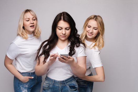 Studio portrait of two girls loking at the mobile phone while blonde girls using it. Gossip or rumour concept. Isolate.