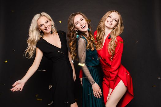 Studio portrait of three elegant stunning women in beautiful evening dresses with makeup posing and smiling at camera. Three beautiful girlfriends celebrating the event together.
