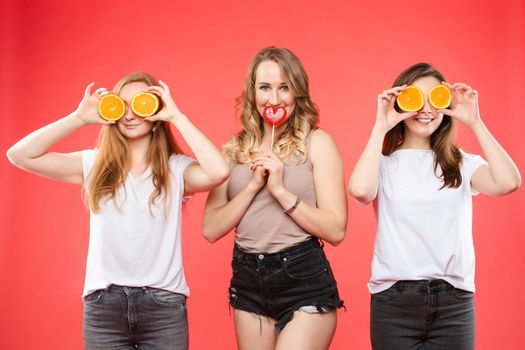 Stock photo of a beautiful girl with a red heart-shaped lollipop between two smiling girls with halved cut oranges. They are isolate on red background.