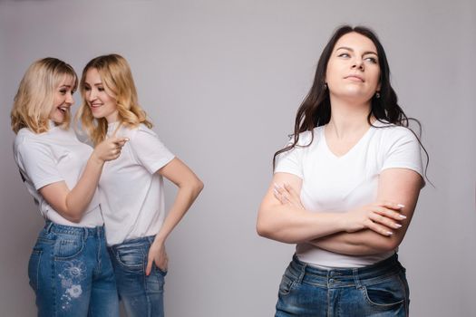 Front view of offended brunette with folded arms looking aside while two blondes standing behind and gossiping. Female friends laughing and talking on grey isolated background. Concept of sneering.