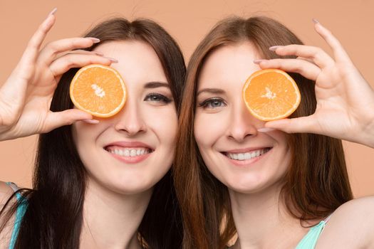 Front view of female friends keeping fresh oranges and posing on isolated background in studio. Beautiful young girls looking at camera and laughing while keeping fruits. Concept of health.