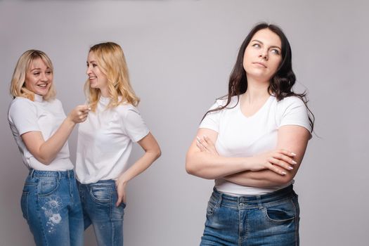 Front view of offended brunette with folded arms looking aside while two blondes standing behind and gossiping. Female friends laughing and talking on grey isolated background. Concept of sneering.