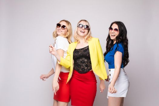 Three cheerful women wearing bright colorful skirts, shirts and glasses standing on grey isolated background. Happy girls looking at camera, laughing and showing tongue. Concept of happiness.