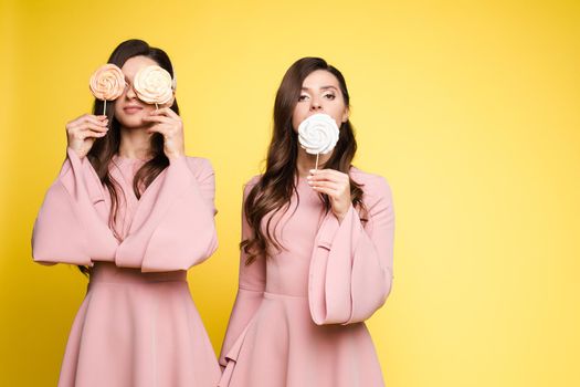 Front view of charming twins in pink dresses closing eyes with lollipops and posing on isolated background. Two female friends laughing and having fun in studio. Concept of childhood and beauty.