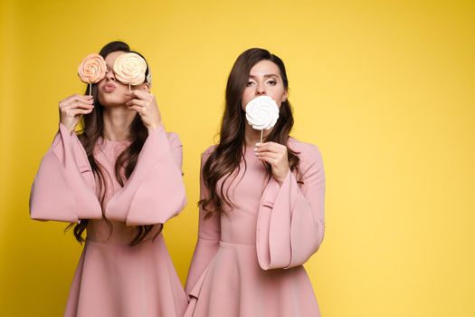 Front view of charming twins in pink dresses closing eyes with lollipops and posing on isolated background. Two female friends laughing and having fun in studio. Concept of childhood and beauty.