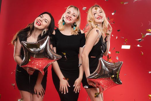 Group smiling beautiful woman posing surrounded by colorful shimmering confetti isolated red studio background. Happy three girl friend enjoying holiday party relaxing and having fun medium long shot
