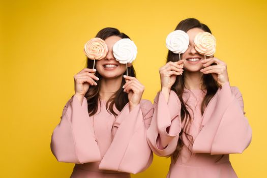 Front view of charming twins in pink dresses closing eyes with lollipops and posing on isolated background. Two female friends laughing and having fun in studio. Concept of childhood and beauty.