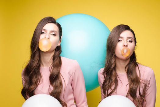 Young twins in elegant dresses holding pink and white balloons in their hands. Brunette sisters with long hair blowing big bubbles from chewing gum. Beautiful girls posing at camera with decorations.