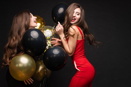 Happy young girls laughing and getting fun together. Brunette beautiful sisters in elegant clothes celebrating birthday. Gorgeous women with red lips standing near bunch of gold and black balloons.