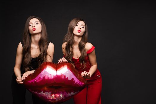 Close up of brunette twins with long hair in black and red costumes posing at camera. Young sisters standing together and holding red lips balloon. Gorgeous models with red lips sending air kisses.