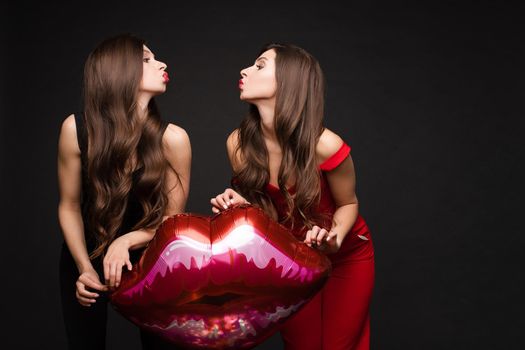 Close up of brunette twins with long hair in black and red costumes posing at camera. Young sisters standing together and holding red lips balloon. Gorgeous models with red lips sending air kisses.