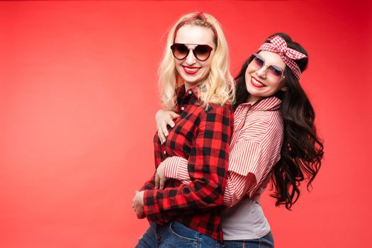 Studio shot of pretty smiling blonde and brunette girlfriends in sunglasses and trendy outlooks hugging against bright red background. Isolated.