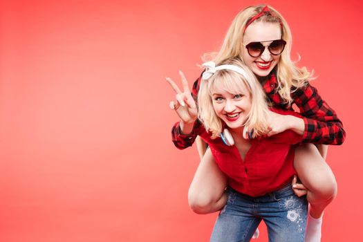 Studio shot of pretty smiling blonde and brunette girlfriends in sunglasses and trendy outlooks hugging against bright red background. Isolated.