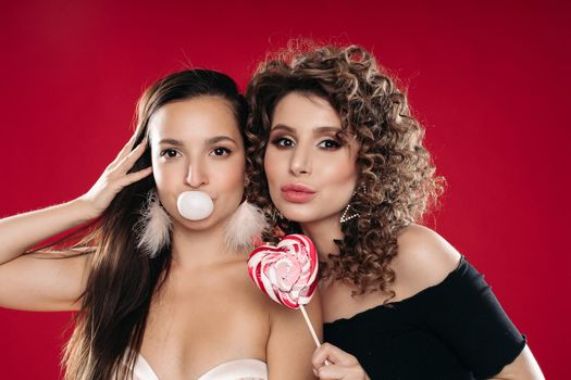 Front view of two different girls looking at camera and posing on black isolated background. Sexy brunette chewing gum while female friend laughing and keeping lollipop in hand. Concept of party.