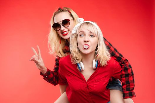 Stock studio portrait of two cheerful and positive trendy girlfriends or sisters having fun. Girl in sunglasses making bunny ears to her friend with her hand while sitting on her back. Isolate on red.