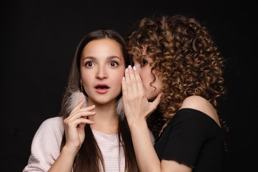 Front view of shocked brunette with big eyes and open mouth in white dress looking at camera while female friend whispering secret on black isolated background. Young women gossiping in studio.