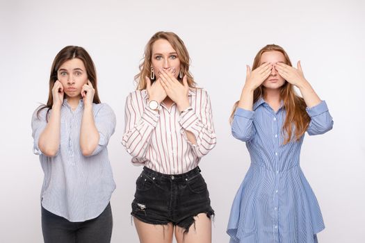 Three young woman friend posing with closing eyes ears and mouth looking at camera medium shot. Stylish female having fun hiding sensory organs isolated at white studio background
