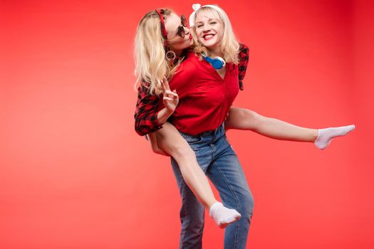 Stock studio portrait of two cheerful and positive trendy girlfriends or sisters having fun. Girl in sunglasses making bunny ears to her friend with her hand while sitting on her back. Isolate on red.