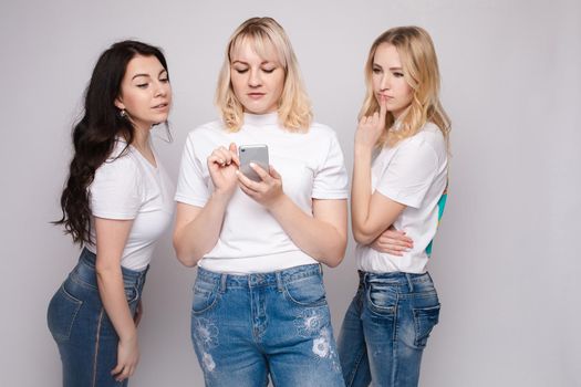 Studio portrait of two girls loking at the mobile phone while blonde girls using it. Gossip or rumour concept. Isolate.