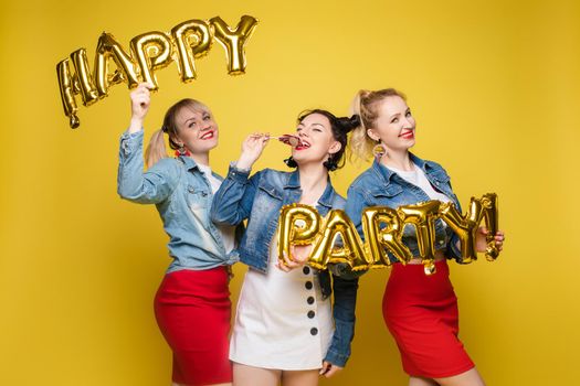 Front view of bright three girls in white shirts keeping balloons and ice creams on yellow isolated background. Cheerful pretty women looking at camera and posing. Concept of party and sparkles.