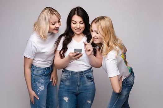 Studio portrait of two girls loking at the mobile phone while blonde girls using it. Gossip or rumour concept. Isolate.