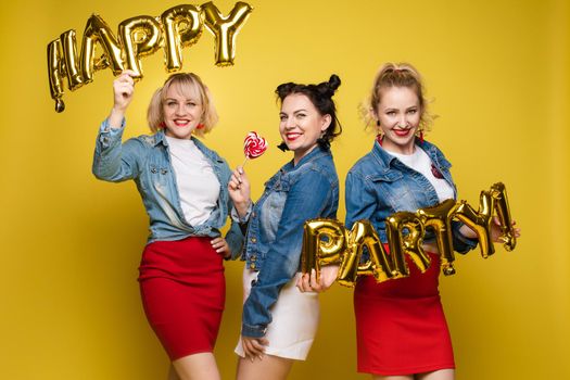 Front view of bright three girls in white shirts keeping balloons and ice creams on yellow isolated background. Cheerful pretty women looking at camera and posing. Concept of party and sparkles.