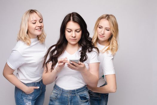Front view of three female friends in white shirt and jeans standing together on grey isolated background and looking at phone. Young blondes and brunette talking and laughing in studio.
